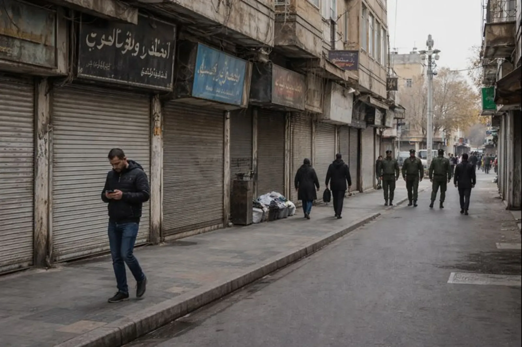 Shuttered shops and light foot traffic on an urban street in Iran with security presence in the distance