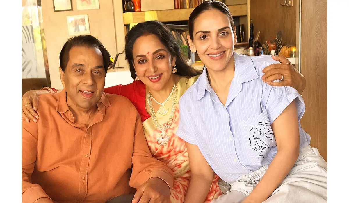 Dharmendra, Hema Malini, and Esha Deol smiling together in a cozy family photo taken indoors.